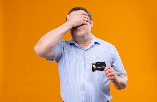 Смущенный и напряженный мужчина среднего возраста в синей полосатой рубашке держит кредитную карту, прикрывая глаза рукой