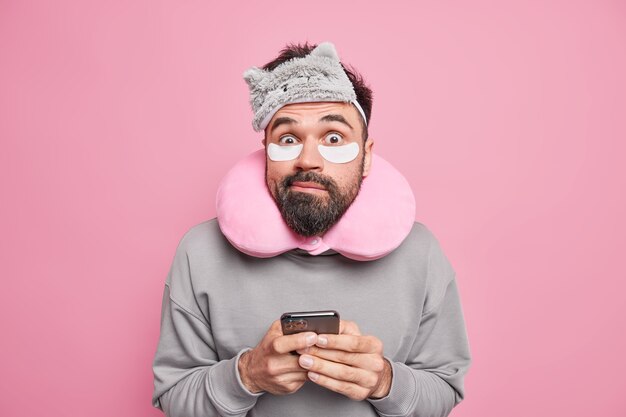 Смущенный удивленный мужчина с бородой использует подушку для шеи для лучшего сна, накладывает косметические патчи под глаза, держит позы для смартфона в помещении у розовой стены, избегает скованности, собираясь вздремнуть