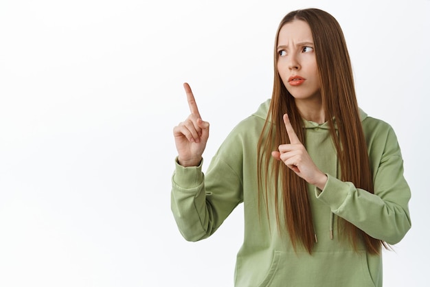 Смущенная девочка-подросток с длинными волосами, указывающая и отводящая взгляд влево, хмурится, озадаченно читает логотип, стоящий на белом фоне