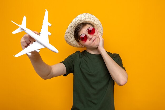 Confused tilting head putting hand on cheek young handsome guy wearing hat with glasses holding toy airplane 