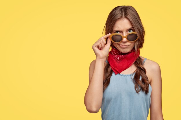 Confused uncertain Caucasian woman keeps hand on sunglasses, raises eyebrows, wears red bandana, has slightly combed plaits, looks with gloomy expression upwards, isolated over yellow  wall