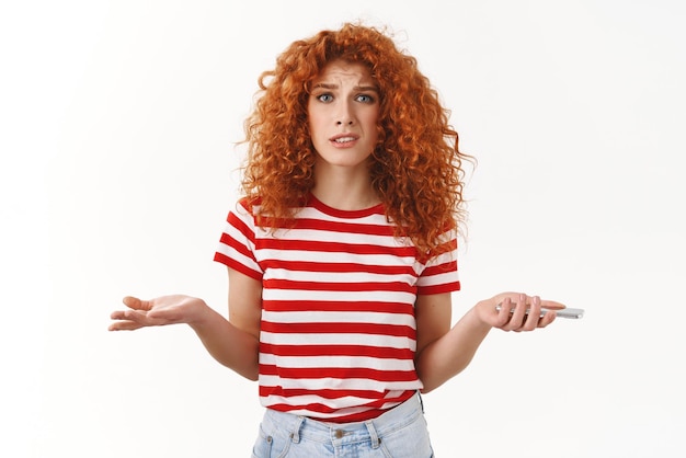 Confused uncertain stylish beautiful redhead curly girlfriend shrugging dismay puzzled holding smartphone hands spread sideways stare questioned trying understand who spam mailbox