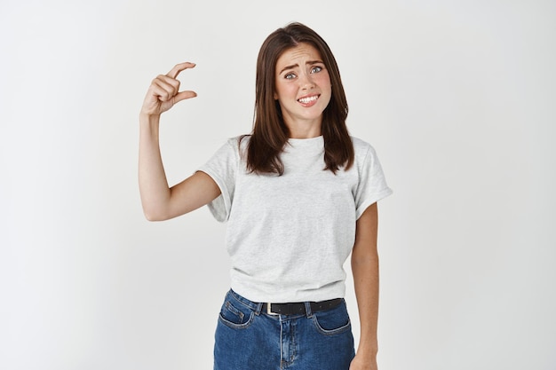 Confused and unhappy young woman showing small size gesture, shaping something tiny and complaining, looking awkward at front.