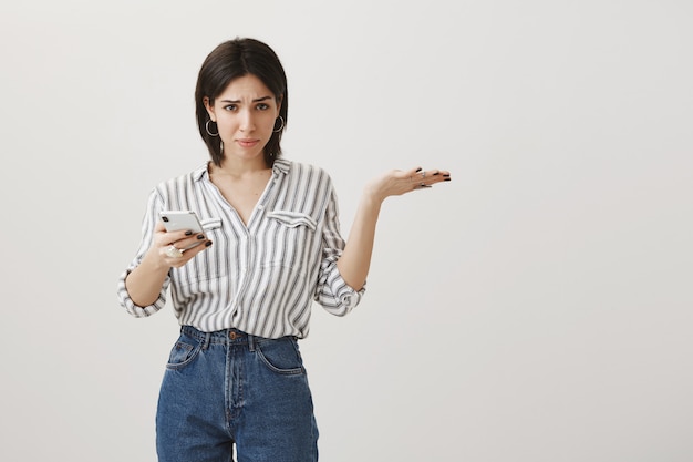 Confused woman asking what while using mobile phone, shrugging puzzled