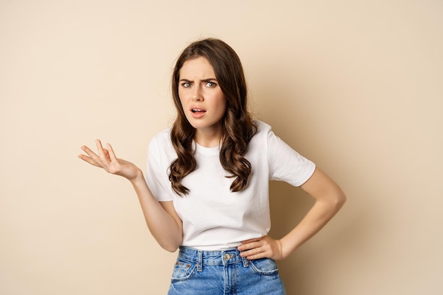Confused woman looking clueless and annoyed, cant understand, asking whats problem, standing over beige background.