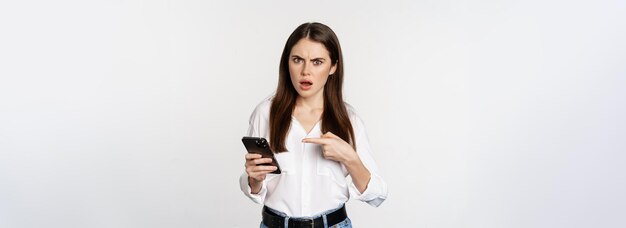 Confused woman pointing at mobile phone and looking puzzled cant figure smth out standing over white