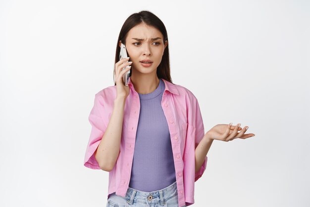 Confused woman talking on mobile phone with puzzled face expression calling on smartphone and shrugging standing over white background