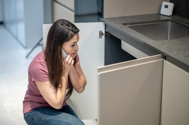 Confused woman with smartphone near ear looking under sink