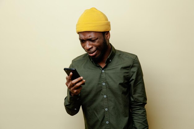 Смущенный молодой африканский американец в шляпе в зеленой рубашке держит и смотрит на телефон, изолированный на белом фоне