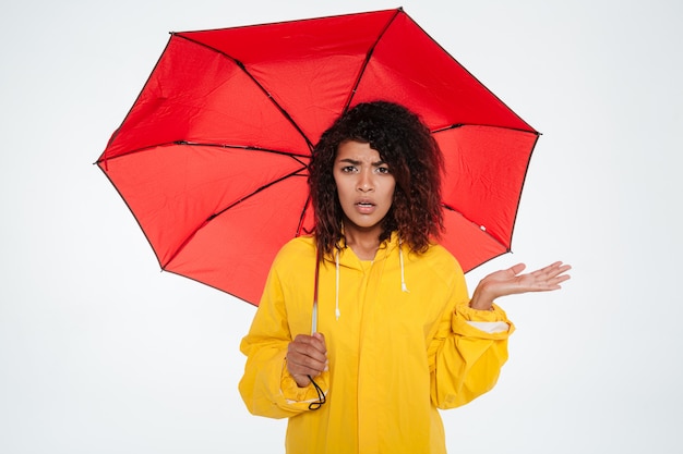 Confused young african woman in raincoat hiding under umbrella