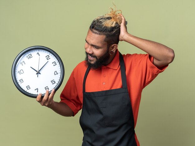 Confused young afro-american barber wearing uniform holding and looking at clock keeping hand on head