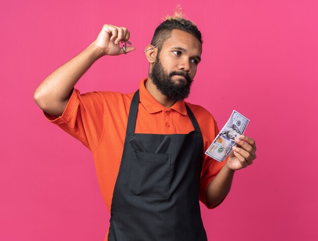 Confused young afro-american male barber wearing uniform raising scissors and holding dollar looking at side