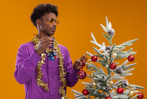 Confused young afro-american man wearing glasses with tinsel garland around neck standing near decorated christmas tree holding christmas baubles looking at tree isolated on orange wall
