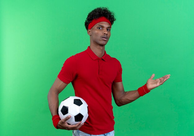 Confused young afro-american sporty man wearing headband and wristband holding ball and points with hand to side isolated on green background with copy space