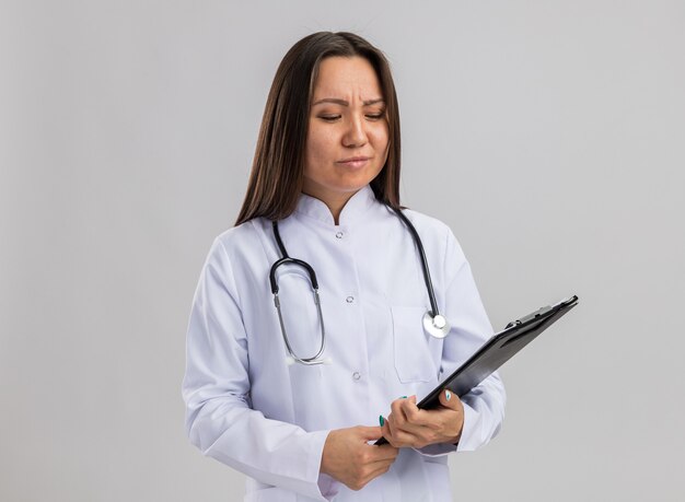 의료 가운과 청진기를 착용하고 복사 공간이 있는 흰색 벽에 격리된 클립보드를 보고 있는 혼란스러운 젊은 아시아 여성 의사