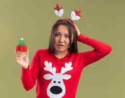 Free photo confused young asian girl wearing christmas hair hoop holding christmas toy putting hand on head isolated on olive green background