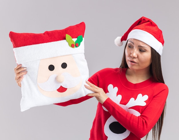 Free photo confused young asian girl wearing christmas hat with sweater holding and looking at christmas pillow isolated on white background