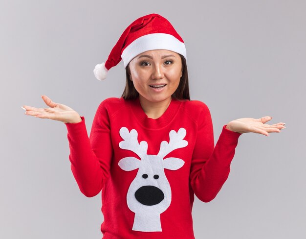 Confused young asian girl wearing christmas hat with sweater spreading hands isolated on white background
