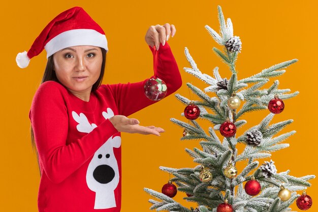 오렌지 벽에 고립 된 크리스마스 트리 볼을 들고 크리스마스 트리 근처에 서 스웨터와 크리스마스 모자를 쓰고 혼란 된 젊은 아시아 여자