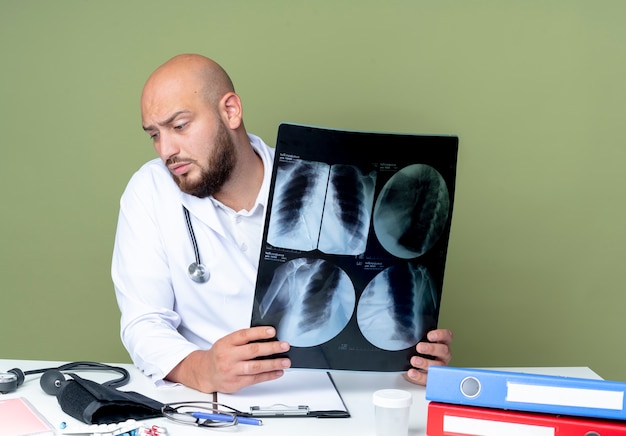 Confused young bald male doctor wearing medical robe and stethoscope sitting at desk work with medical tools holding x-ray isolated on green background