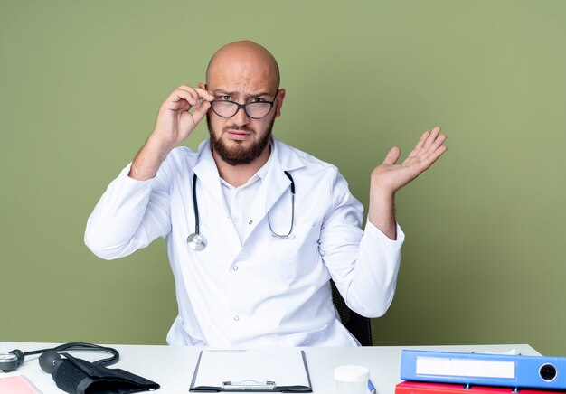 Confused young bald male doctor wearing medical robe and stethoscope sitting at desk work with medical tools take on glasses and spread hand isolated on green background
