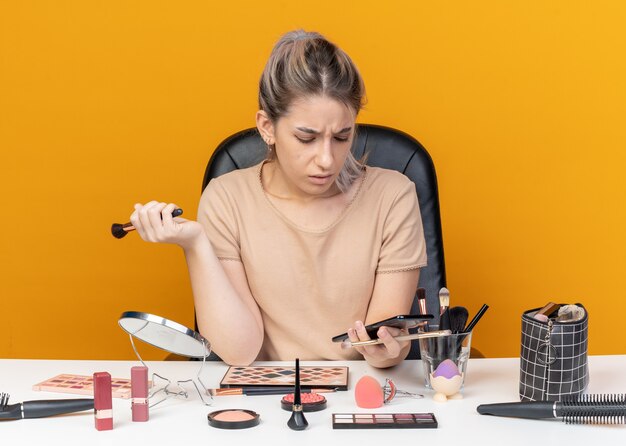 Confused young beautiful girl sits at table with makeup tools holding makeup brush and looking at phone in her hand isolated on orange wall