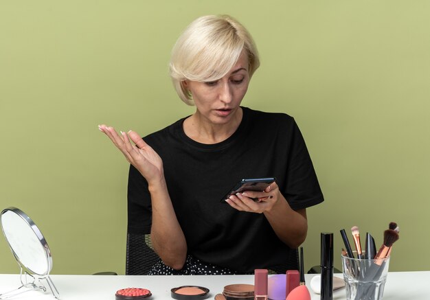 Foto gratuita la giovane bella ragazza confusa si siede al tavolo con gli strumenti per il trucco guardando il telefono in mano isolato sul muro verde oliva