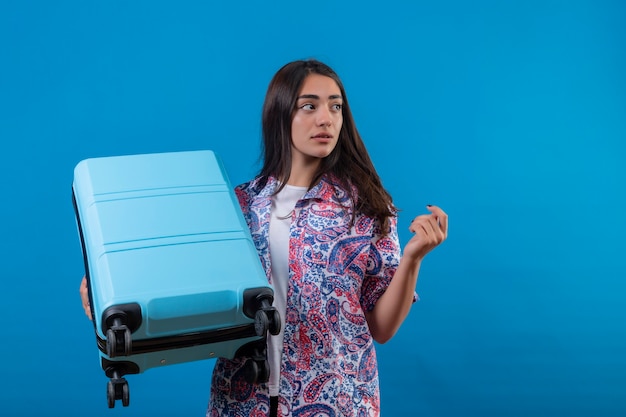 Confused young beautiful traveler woman holding travel suitcase looking aside with doubtful facial expression standing over isolated blue space