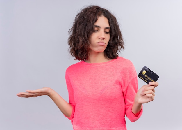 Confused young beautiful woman holding card and showing empty hand on isolated white wall