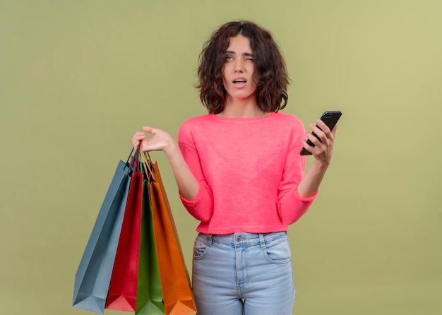 Giovane bella donna confusa che tiene i sacchetti di cartone e il telefono cellulare sulla parete verde isolata