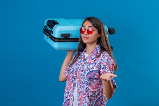 Free photo confused young beautiful woman tourist wearing red sunglasses holding travel suitcase  doubtful shrugging shoulders standing over isolated blue space