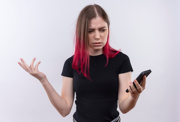 Confused young beautiful woman wearing black t-shirt looking at phone on her hand on isolated white wall