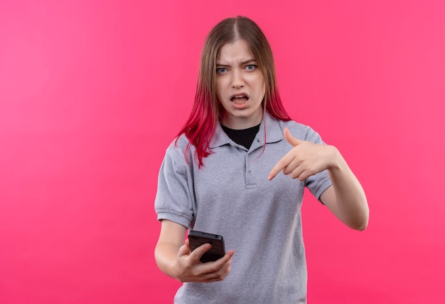 격리 된 분홍색 벽에 그녀의 손에 전화 회색 티셔츠 포인트를 입고 혼란 젊은 아름 다운 여자