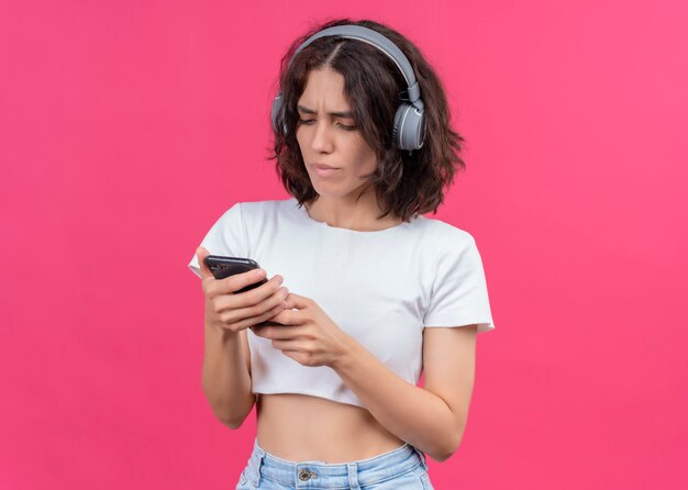 Confused young beautiful woman wearing headphones and holding mobile phone on pink wall with copy space