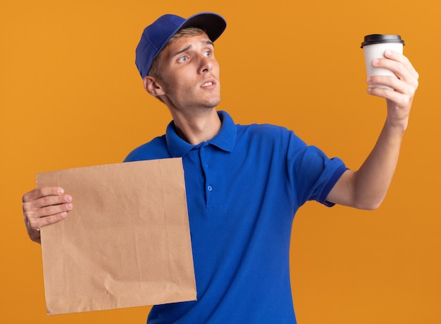 Free photo confused young blonde delivery boy holds paper package and looks at cup on orange