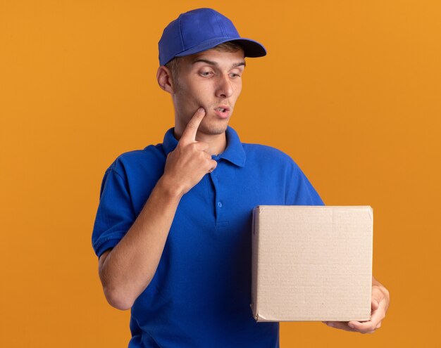 Confused young blonde delivery boy puts finger on chin holding and looking at cardbox