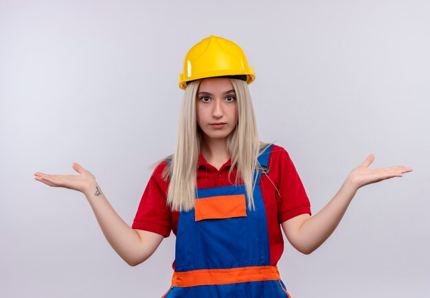 Confused young blonde engineer builder girl in uniform showing empty hands on isolated white space