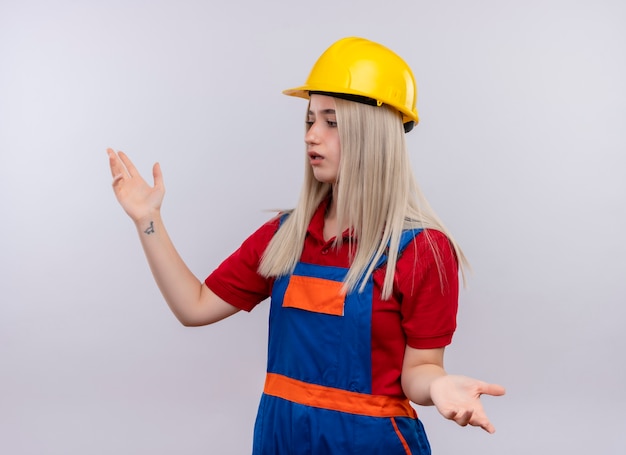 Confusa giovane ragazza bionda ingegnere costruttore in uniforme con le braccia aperte guardando il lato sinistro su uno spazio bianco isolato