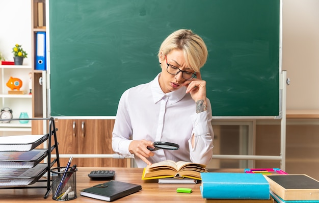 Запутанная молодая блондинка учительница в очках сидит за столом со школьными принадлежностями в классе, глядя на открытую книгу через увеличительное стекло, касаясь головы