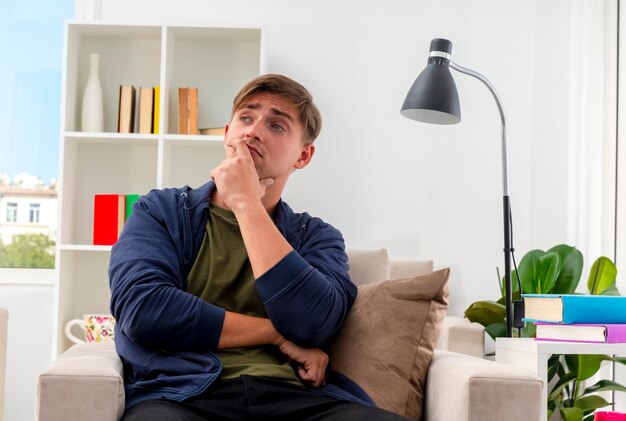 Confused young blonde handsome man sits on armchair putting hand on chin looking at side inside the living room