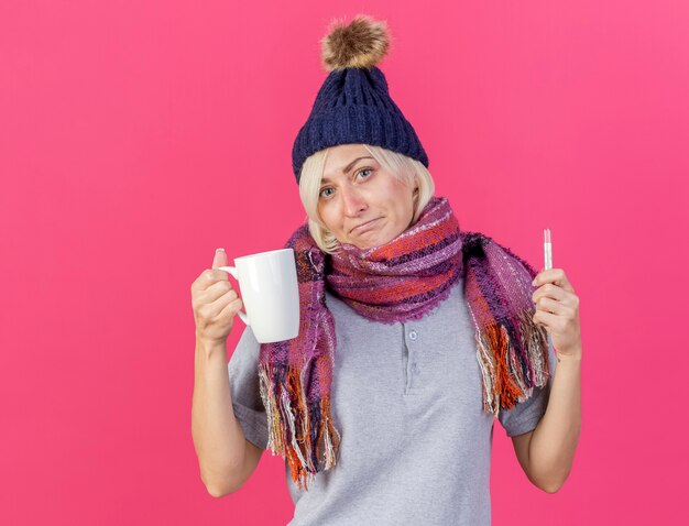 Confused young blonde ill slavic woman wearing winter hat and scarf holds cup