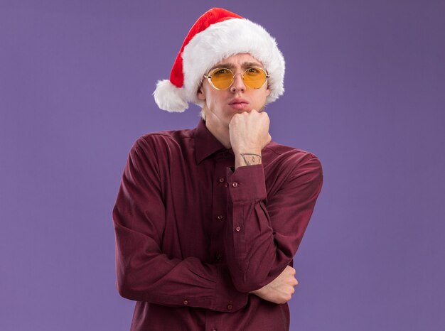 Confused young blonde man wearing santa hat and glasses looking at camera keeping hand on chin isolated on purple background with copy space