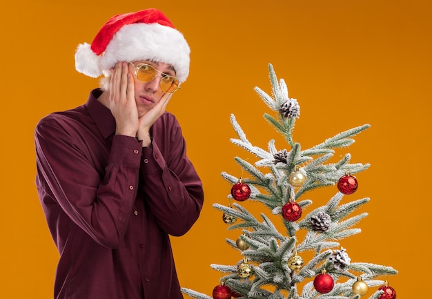 Confused young blonde man wearing santa hat and glasses standing near decorated christmas tree on orange background