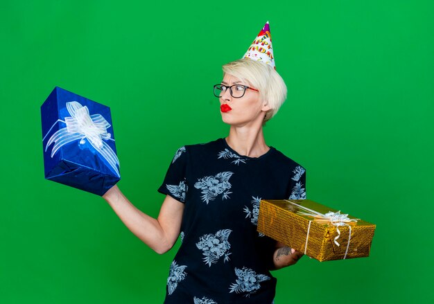 Confused young blonde party girl wearing glasses and birthday cap holding gift boxes looking at one of them isolated on green background