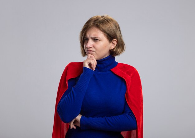 Confused young blonde superhero girl in red cape touching chin looking at side isolated on white background with copy space