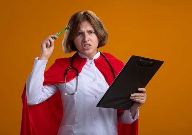 Confusa giovane donna bionda supereroe in mantello rosso che indossa l'uniforme del medico e stetoscopio tenendo appunti e toccando la testa con la matita guardando davanti isolato sulla parete arancione