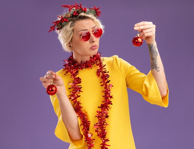 クリスマスの頭の花輪と首の周りに見掛け倒しの花輪を身に着けている混乱した若いブロンドの女性は、紫色の背景に分離されたそれらの1つを見てクリスマスつまらないものを保持している眼鏡