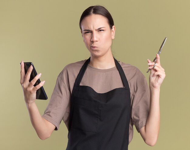 Confused young brunette barber girl in uniform holds hair thinning scissors and phone