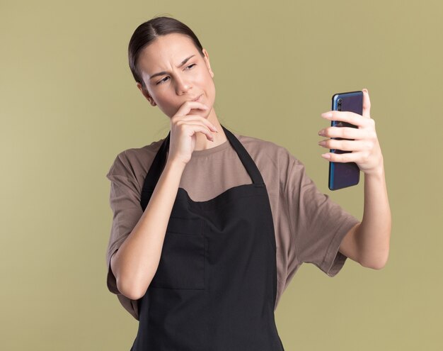 Смущенная молодая брюнетка-парикмахер в униформе кладет руку на подбородок, держа и глядя в телефон на оливково-зеленом