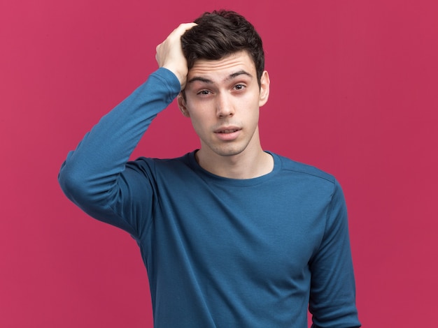 Free photo confused young brunette caucasian boy puts hand on head looking at camera on pink